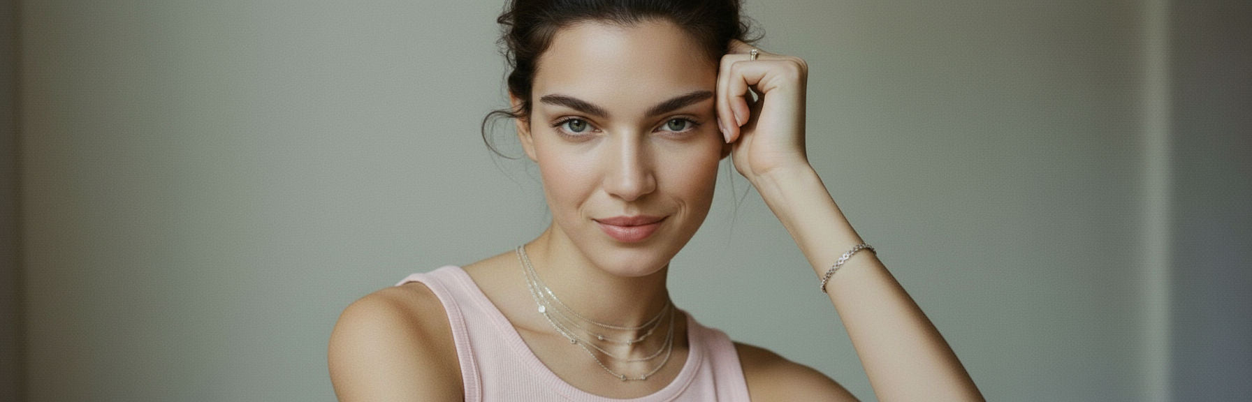 Woman in pink tank top wearing layered silver necklaces, a silver bracelet, and a ring, hand near her face.