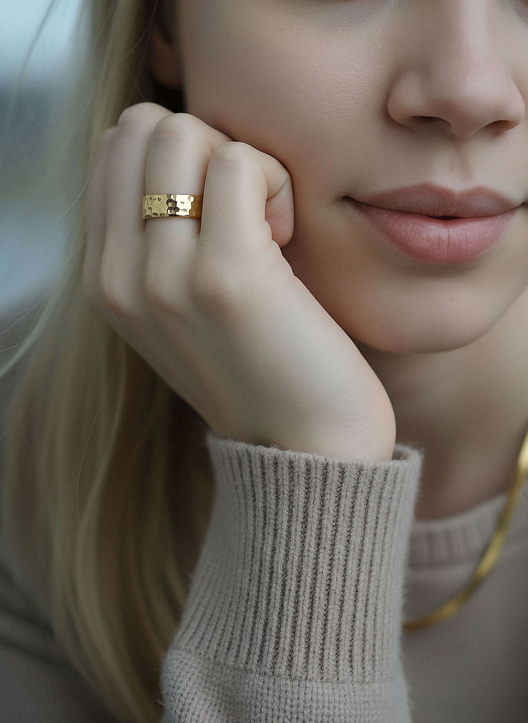 Blonde woman wearing a wide, hammered gold ring on her right index finger, resting her chin on her hand.